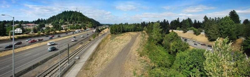 A landscape featuring a highway with cars, railway tracks, trees, and a distant hill under a partly cloudy sky.