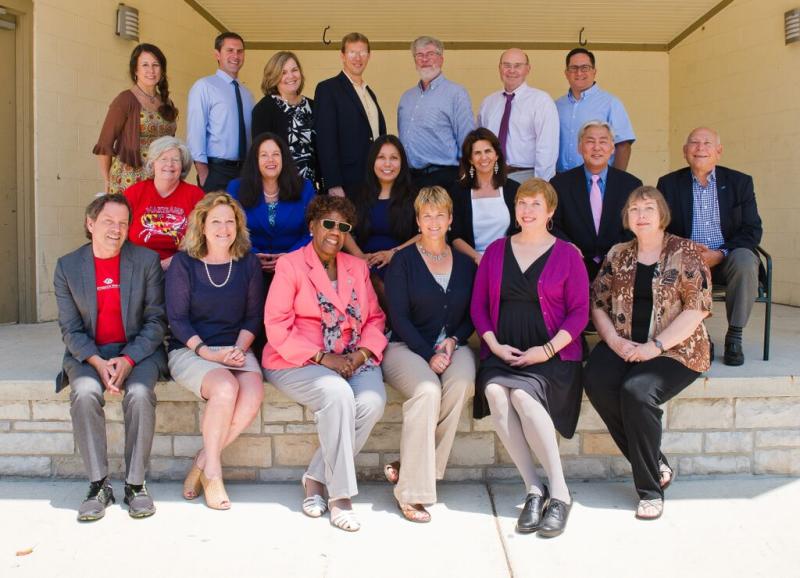 A group of fifteen people pose for a formal photo, two seated rows in front of a light brick wall.