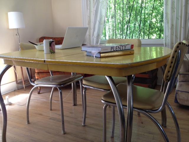 A vintage yellow table with a laptop, coffee mug, and books under a window with white curtains.